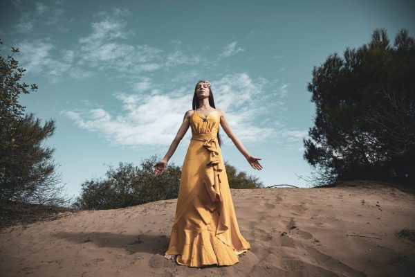 Woman in a flowing yellow dress stands on sandy terrain, arms outstretched, surrounded by trees under a blue sky.