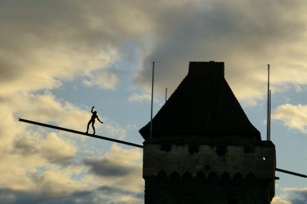 Silhouette of a person balancing on a beam extending from a tall, stone tower against a cloudy evening sky.