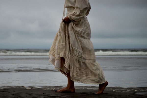 A person in a long, flowing dress walks barefoot along a cloudy beach shoreline.