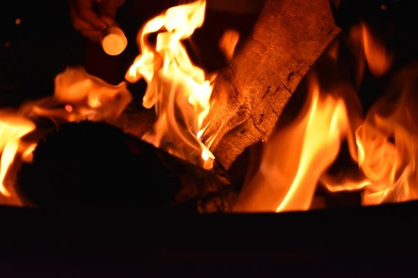 A hand holding a marshmallow over a campfire with bright orange flames and burning logs.
