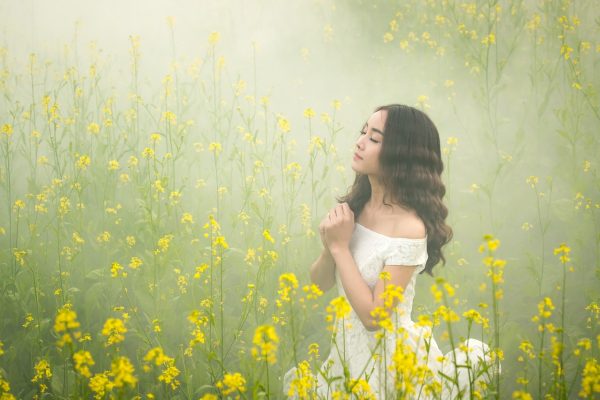 a person in a white dress with long dark wavy hair stands with eyes closed and hands clasped at the heart in the middle of a field of yellow flowers on a foggy day