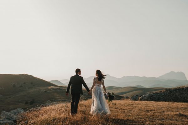 A couple in wedding attire walks hand in hand across a grassy hill with mountains in the background.