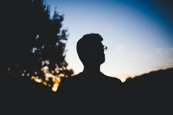 Silhouette of a person with glasses against a sunset sky, surrounded by dark trees.