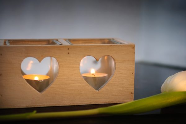 Wooden candle holder with heart cutouts, two lit candles inside, and a white tulip on a dark surface.