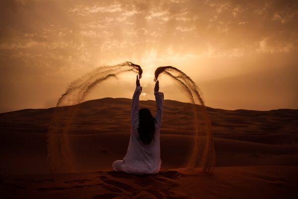 woman with long brown hair shown from behind in a white tunic sitting in desert sand, her arms raised to the sky with arcs of sand falling on both sides, looking at setting sun in grayish brown cloudy sky