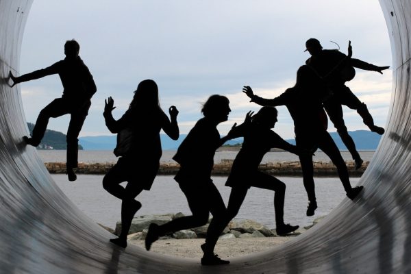 Silhouetted figures in dynamic poses inside a large pipe with a beach and ocean in the background.