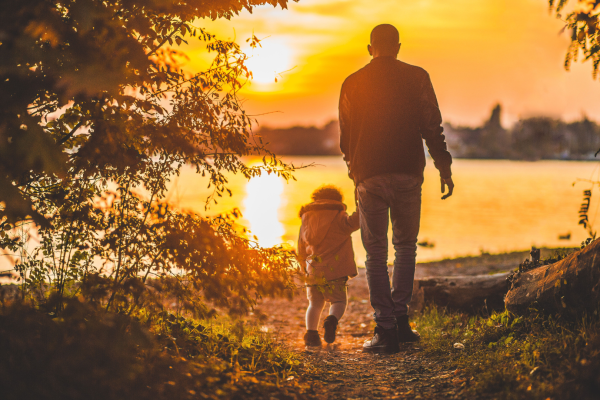 An adult and child walk by a lakeside at sunset, surrounded by trees and warm golden light.