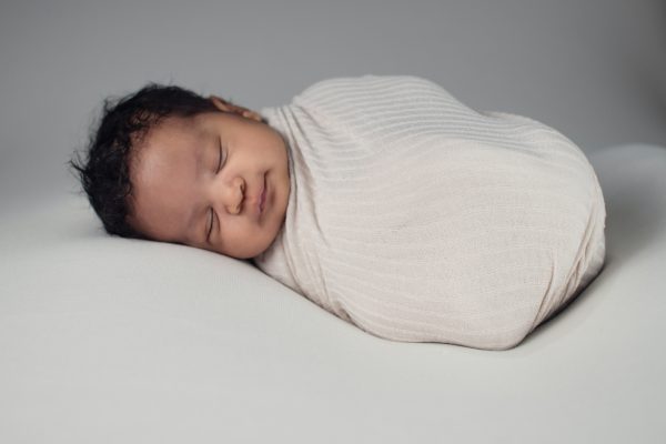 A newborn baby peacefully sleeping, wrapped in a cream-colored blanket against a soft gray background.
