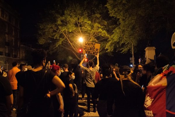 Protesters gather at night, one holding a sign reading White Silence = Violence under a tree.