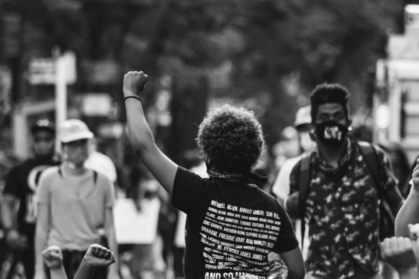 black and white photo of people at a protest, one black/brown woman shown from behind with fist in the air, another black man in mask standing near her and another white person walking by on her other side