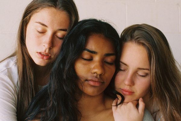 Three women with eyes closed, embracing closely in a serene, intimate moment against a light background.