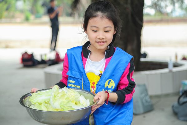 Young girl wearing a blue vest holds a large bowl of leafy lettuce outdoors, smiling.