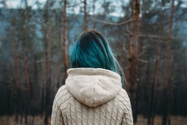 Person with teal hair in a beige sweater facing a forest in winter.