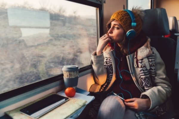 A woman with headphones gazes out a train window, with a coffee cup, orange, and tablet on the table in front of her.