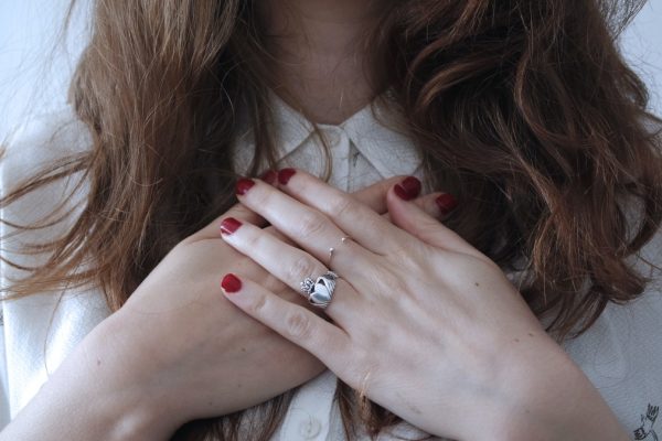 Woman with red nail polish holding hands on chest, wearing a silver ring and a white shirt.