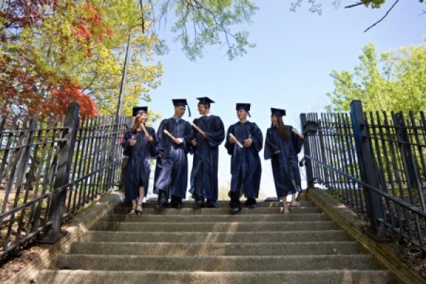 Five graduates in caps and gowns walk down steps outdoors, surrounded by trees and fence.