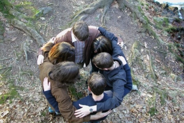 A group of people standing in a circle with arms around each other in a wooded area.