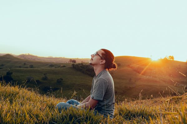 Person sitting on grassy hill at sunset, looking up at the sky, surrounded by rolling hills.
