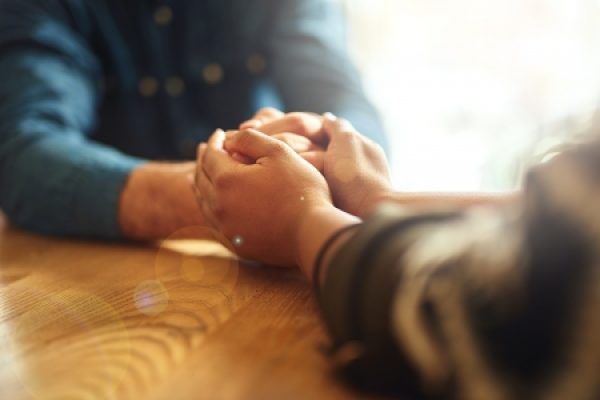 Two people seated at a wooden table, holding hands in a comforting manner with sunlight coming from the background.