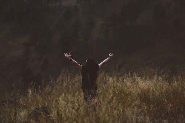 Person standing in a field with arms raised, surrounded by tall grass and trees in the background.