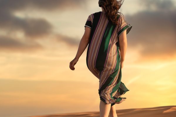 woman walking barefoot in desert wearing loose striped dress with hair blowing in the wind