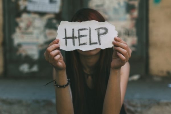 Person holding a torn paper with HELP written on it, covering their face, in an urban setting.