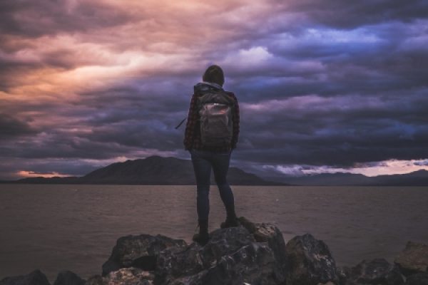 Person with a backpack stands on rocks overlooking a large body of water under a dramatic, cloudy sunset sky.
