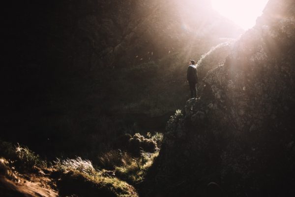 Person standing on a rocky ledge, bathed in sunlight, surrounded by dark cliffs and greenery.