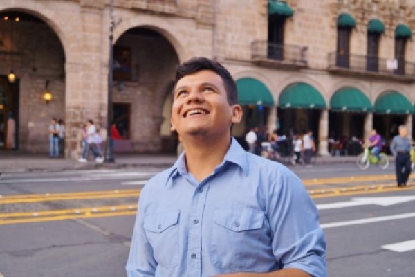 A smiling man in a blue shirt looks upward on a city street with historic buildings in the background.