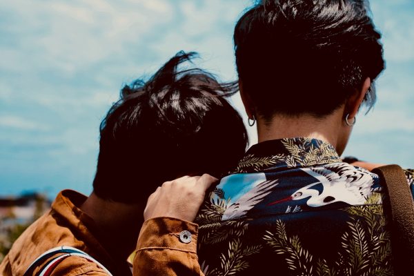 Two people with dark hair stand together closely outdoors, one with a printed shirt featuring birds and foliage.