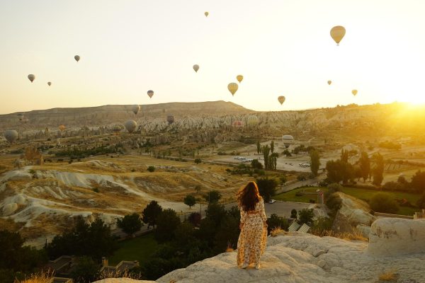Woman in a floral dress watching hot air balloons at sunrise over a rocky landscape.