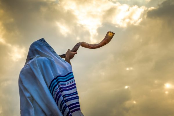 Person blowing a shofar, wearing a tallit against a dramatic cloudy sky.