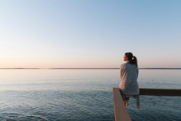 Person sitting on a railing, gazing at a calm sea during sunset.