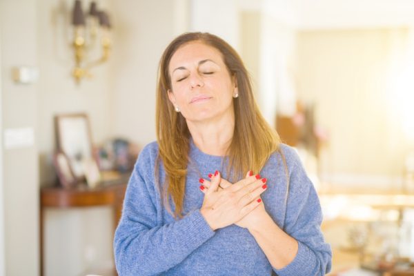 A woman with closed eyes holds her hands to her chest, expressing serene gratitude, in a brightly lit room.