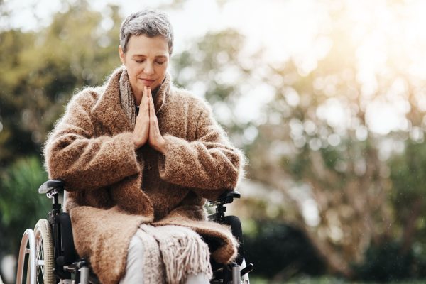 A person in a wheelchair, wearing a cozy coat, peacefully meditates with hands together outdoors in sunlight.