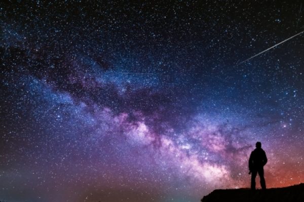 A person stands on a hill, gazing at the star-filled night sky and the Milky Way galaxy, with a shooting star above.