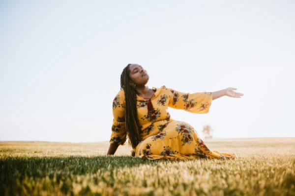 a person with long hair and a yellow flowing dress sits in a field with closed eyes and an outstretched arm