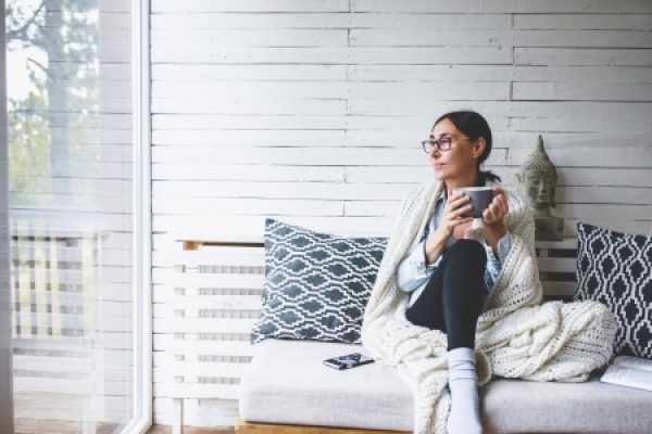 a person wearing glasses, a sweater and leggings holds a coffee cup and sits on a couch, looking to the side