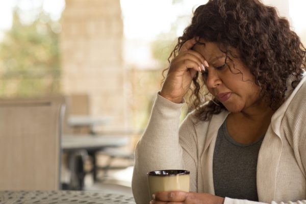 A woman sits at an outdoor table, holding a mug, looking contemplative.