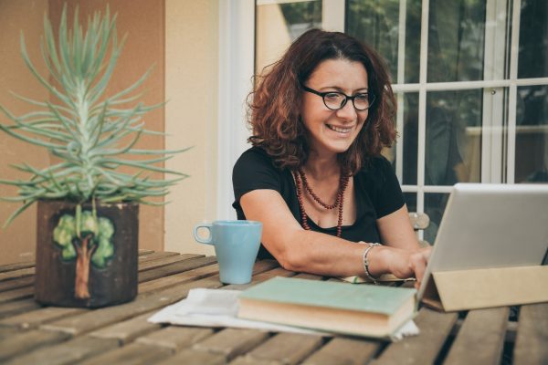 Smiling woman with glasses using a tablet at an outdoor table, next to a plant, cup, and books.
