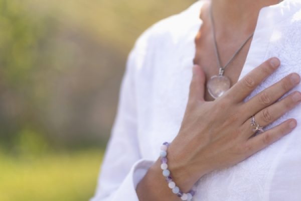 A person wearing a white top holds their hand to their chest, showing jewelry and a beaded bracelet.