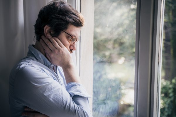 Man in glasses looking pensive by a window, holding his face with one hand, wearing a light blue shirt.