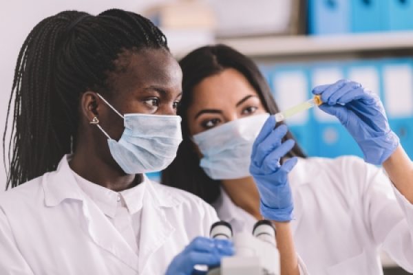 Two scientists in lab coats and masks examine a test tube near a microscope.