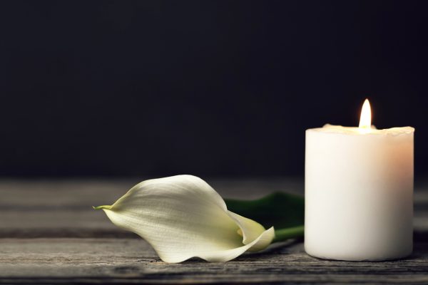 lit candle, thick and white, next to white petal on wood table against black background