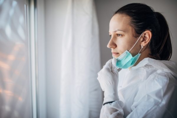 Person in protective clothing and mask looking out a window, appearing thoughtful.