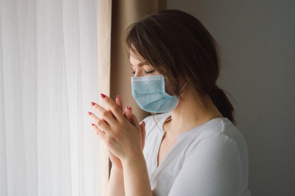 Woman wearing a mask standing by a window, eyes closed, hands clasped together.