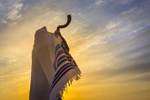 Person wrapped in a prayer shawl blowing a shofar against a sunset sky.