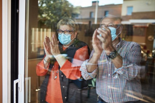 Two elderly people wearing masks applaud inside while looking out of a window.