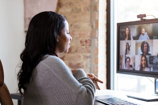 brown skinned woman with dark hair sitting at desk looking at computer screen where an array of faces are shown as on a Zoom screen