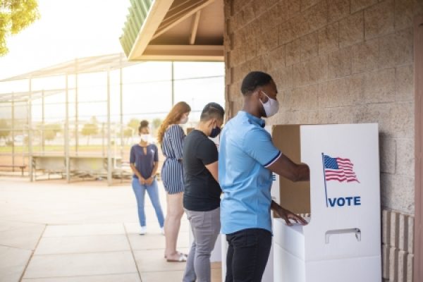 People wearing masks, waiting and casting votes in outdoor polling booths with U.S. flag.